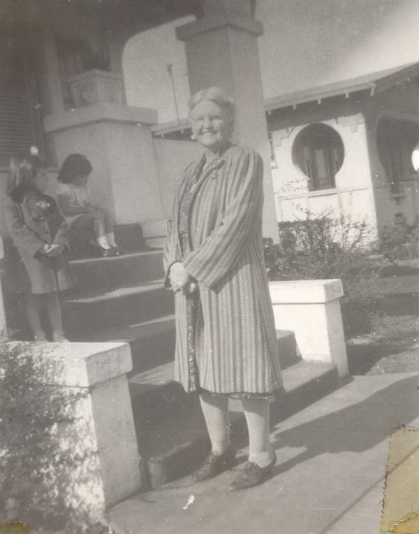 Bertha in a long coat at the bottom of the front steps of a house
